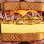Korean street toast cut diagonally showing steamed egg patty, cabbage fritter, ham, cheese, and sauces