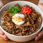 Gyudon in a white bowl, thin-sliced beef and browned onions over rice, topped with an onsen egg, beni shoga, and scallions
