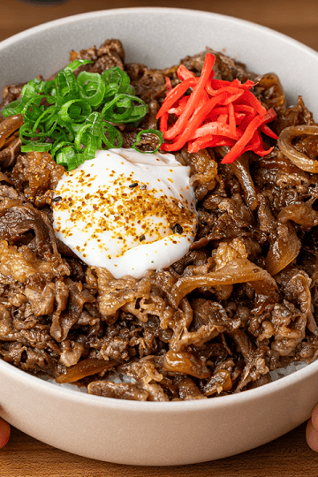 Gyudon in a white bowl, thin-sliced beef and browned onions over rice, topped with an onsen egg, beni shoga, and scallions