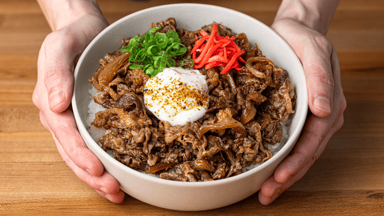 Gyudon in a white bowl, thin-sliced beef and browned onions over rice, topped with an onsen egg, beni shoga, and scallions