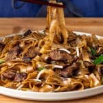Chopsticks lifting wide rice noodles and seared beef from a plate of dry-fried beef chow fun
