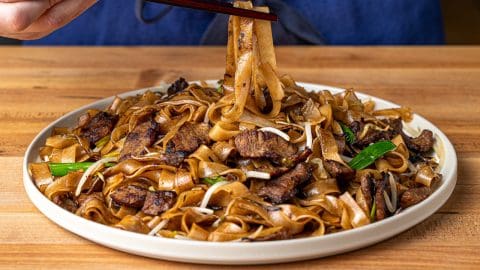 Chopsticks lifting wide rice noodles and seared beef from a plate of dry-fried beef chow fun