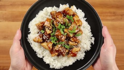 Teriyaki chicken pieces with sesame seeds and sliced scallions over white rice on a black plate held in two hands