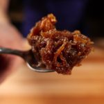 A spoonful of dark brown caramelized onions held up close over a wooden cutting board