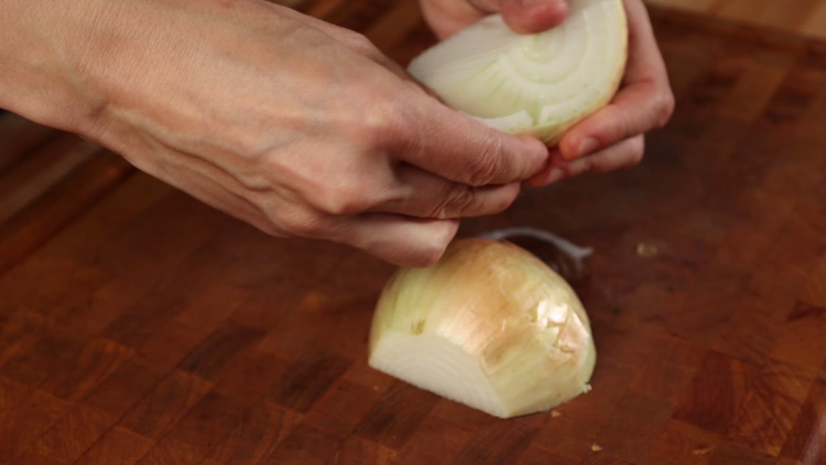 Hands peeling the papery skin off a halved yellow onion on a wooden cutting board