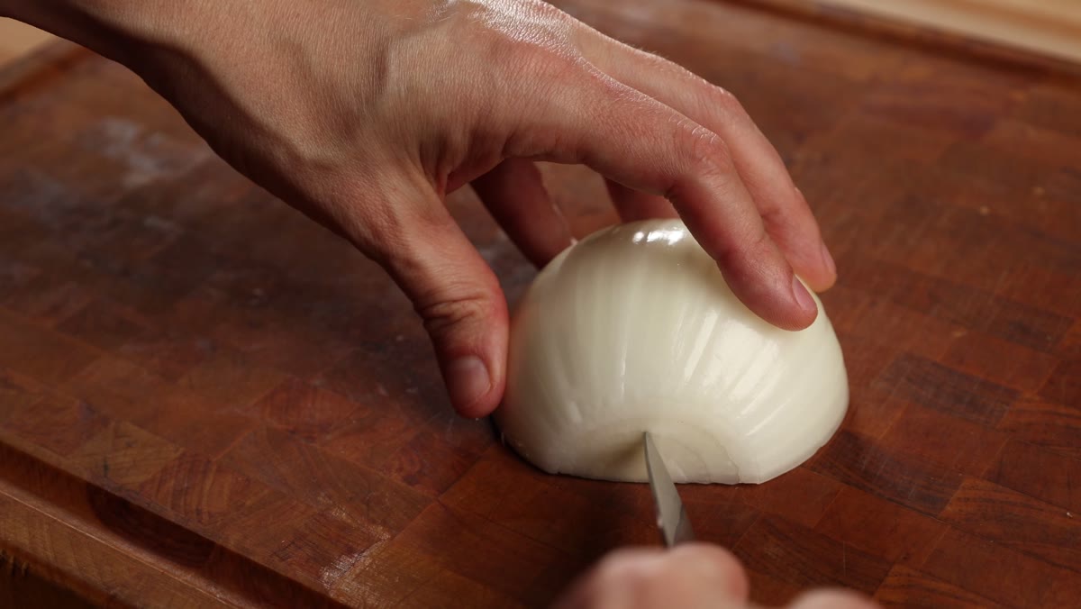 A paring knife making a small V-shaped cut to remove the root from a peeled onion half
