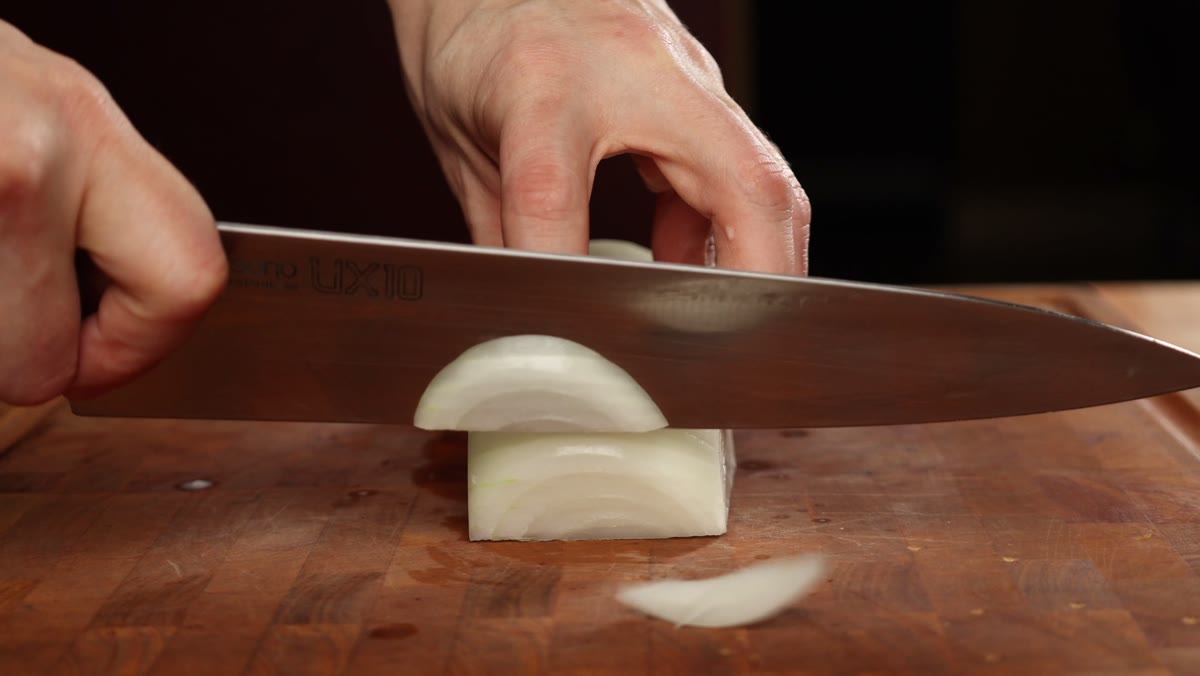 A chef's knife slicing a peeled onion half into thin strips along the grain