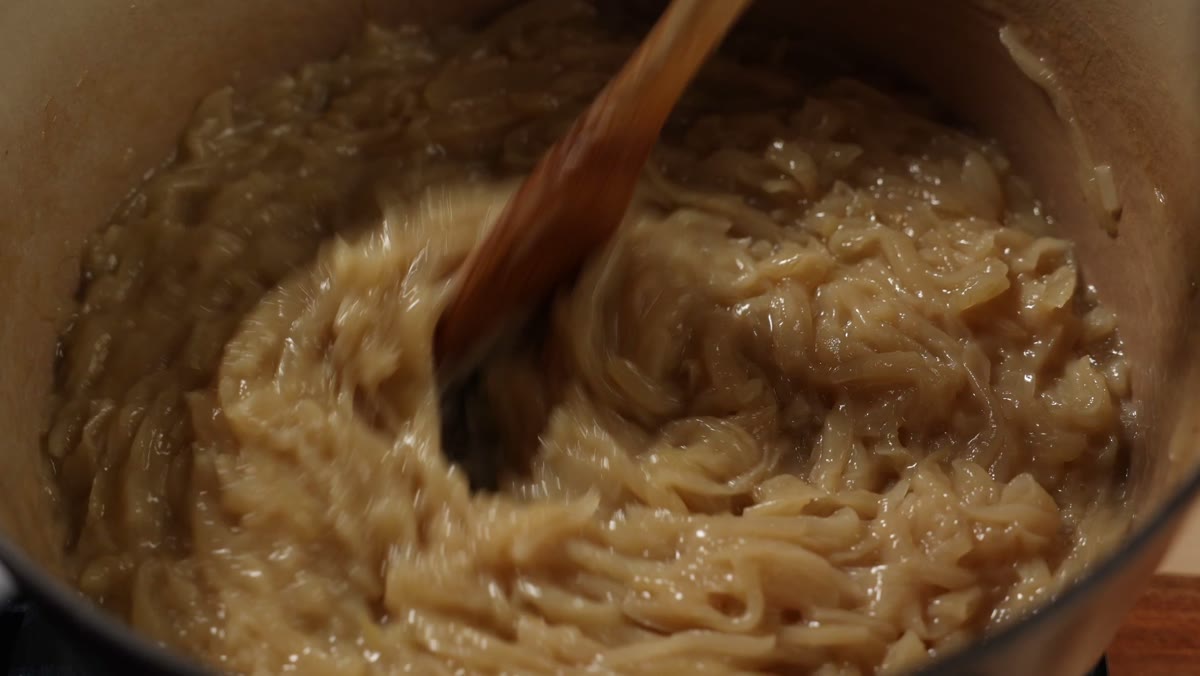 A wooden spoon dragged through soft golden onions to check for liquid on the bottom of the pot