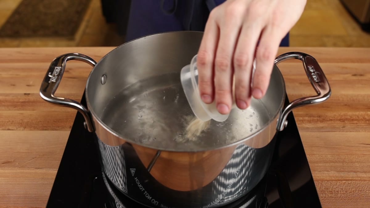 Chicken bouillon powder being poured from a small cup into a pot of simmering water