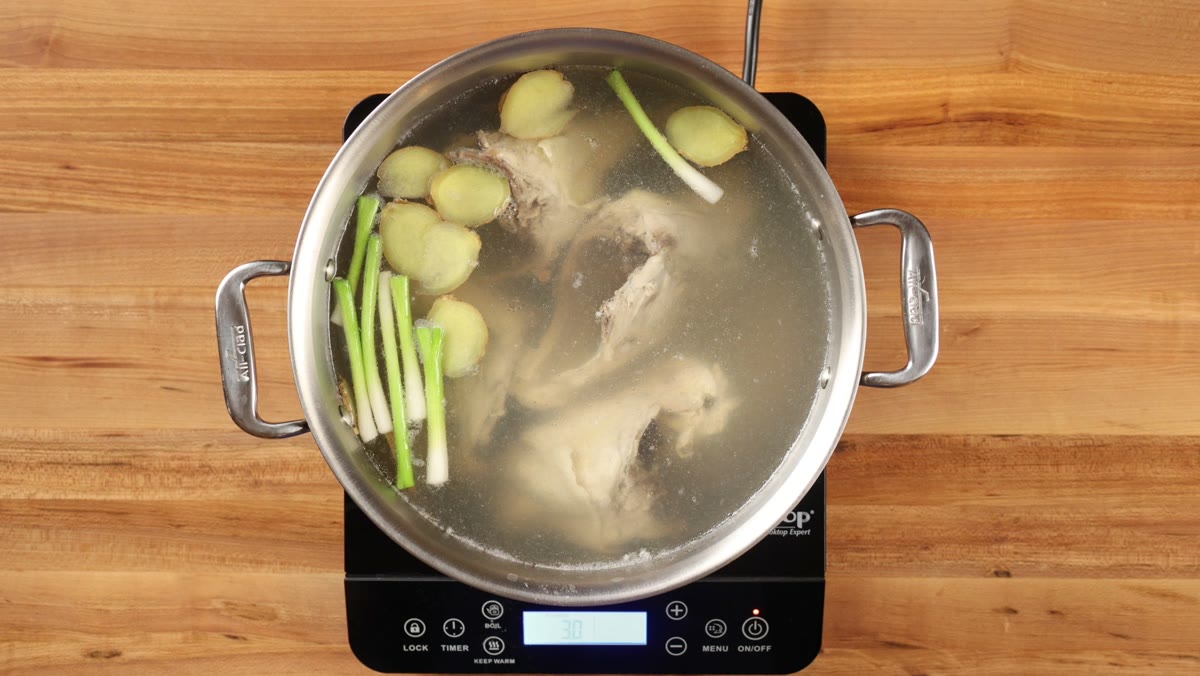 Chicken carcasses simmering with ginger and green onions in a large pot, seen from above