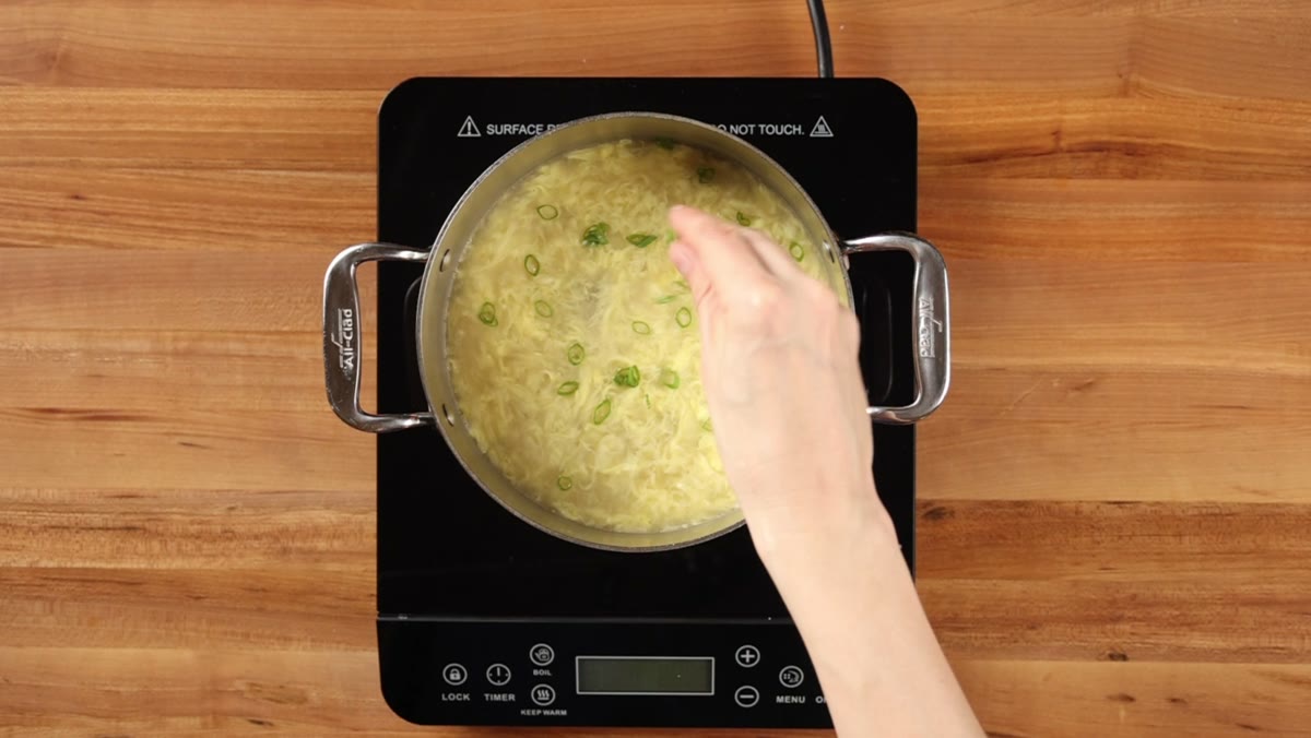 A hand stirring sliced green onions into finished egg drop soup in the pot, seen from above