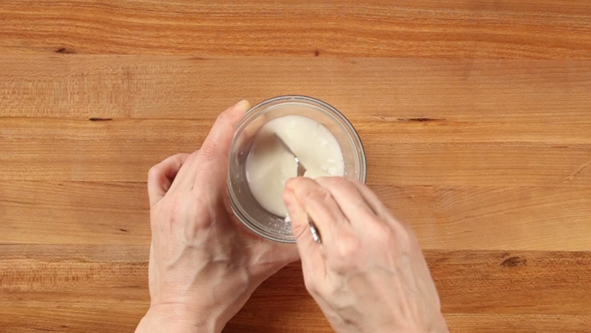 Hands stirring cornstarch and cold water into a slurry in a small glass bowl