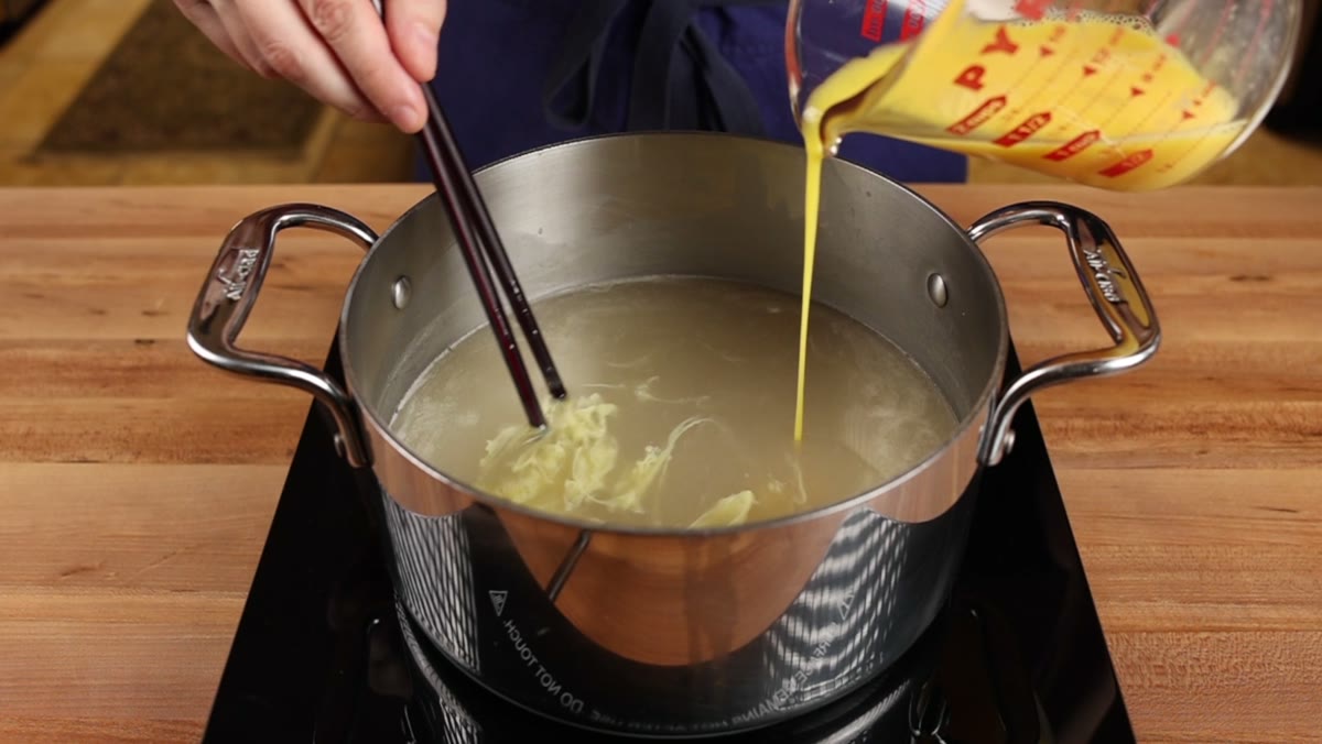 A thin stream of beaten egg poured into the broth while chopsticks stir the pot