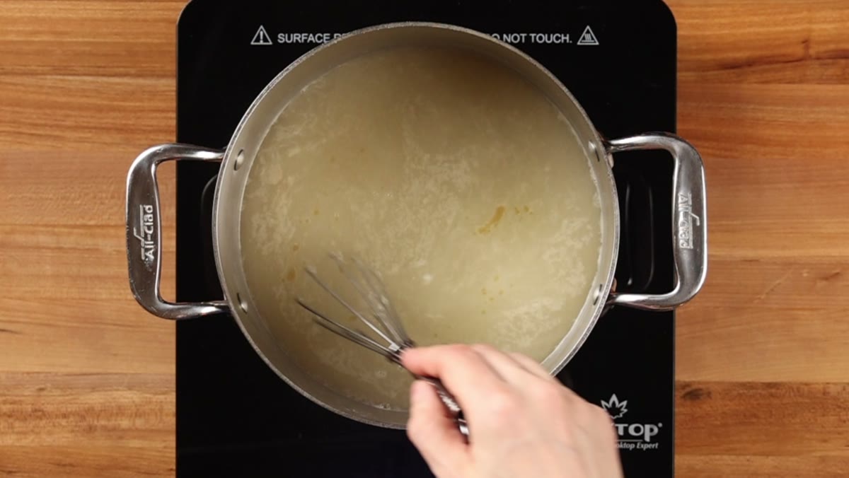A whisk stirring the seasonings into chicken broth in a steel pot, seen from above