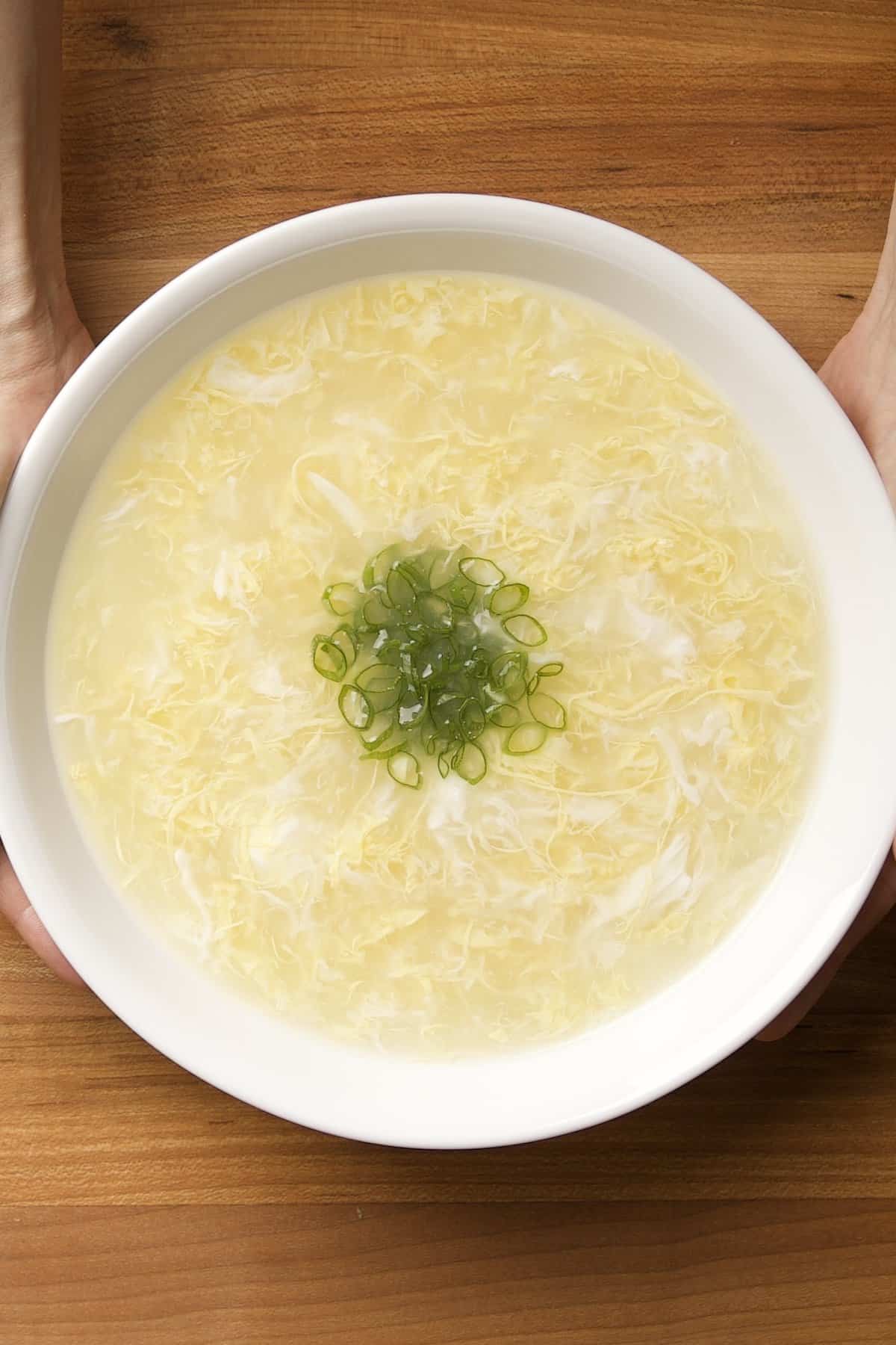 A white bowl of egg drop soup with wispy egg ribbons and sliced green onions on a wooden table