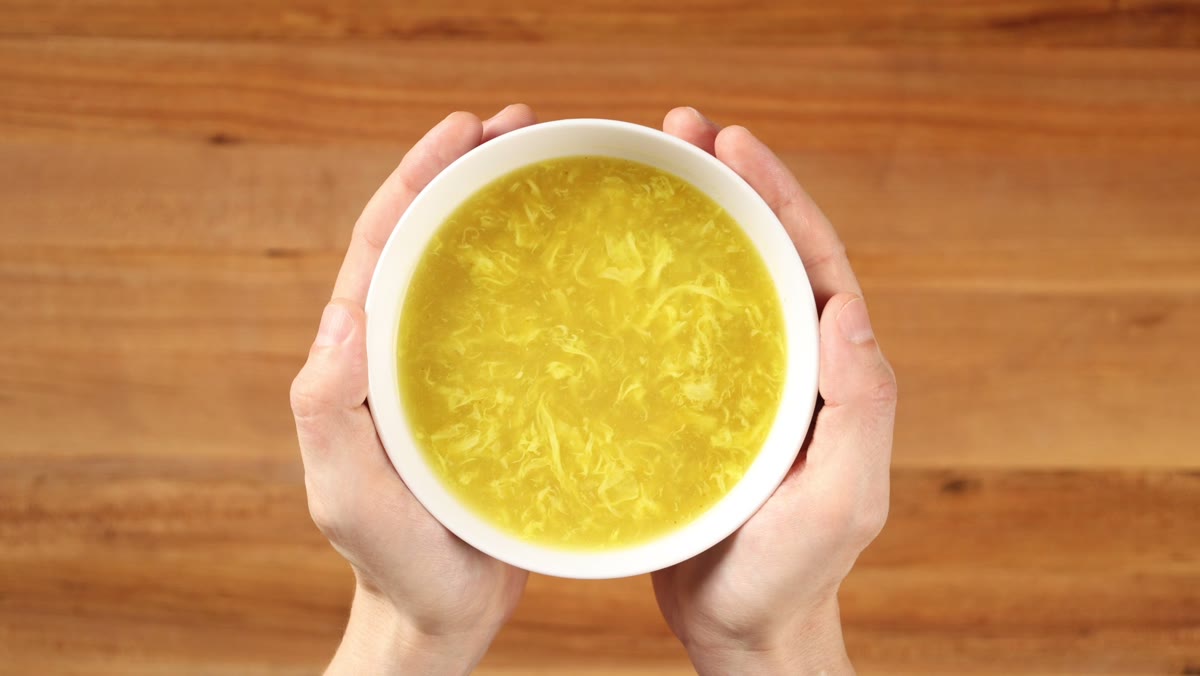 Two hands holding a bowl of bright yellow egg drop soup colored with turmeric