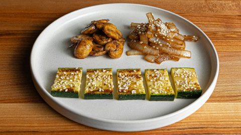Seared mushrooms and sesame-topped onions above a row of browned zucchini pieces on a white plate