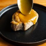 A spoonful of golden miso sauce poured over a pork chop on a black plate